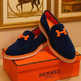 Navy loafers with orange accents on an Hermès box, with Hermès branding in the background.
